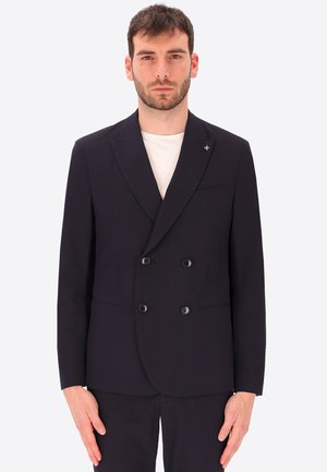Mann trägt einen schwarzen zweireihigen Blazer über einem weißen Hemd, steht vor einem schlichten hellen Hintergrund.
