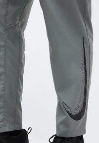 Γκρι παντελόνι με μεγάλο μαύρο λογότυπο Nike swoosh στο κάτω μέρος του ποδιού, φορεμένο με μαύρα αθλητικά παπούτσια σε ανοιχτό φόντο.