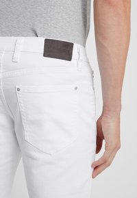 Vita denimjeans med en texturerad yta, med en brun läderetikett, bakfickor och silverfärgade metalldetaljer.