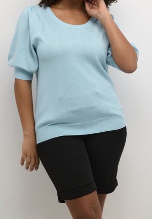 Kobieta w jasnoniebieskim swetrze z krótkimi bufiastymi rękawami i czarnych spodenkach do kolan z podwiniętymi brzegami, pozująca na tle jednolitego tła.