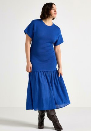 Kvinde iført en blå strikbluse med korte ærmer, en plisseret blå midi-nederdel og mørkebrune knæhøje støvler, stående foran en ensfarvet baggrund.