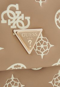 Détail de sac beige avec plaque logo Guess triangulaire dorée et motifs floraux et lettres blancs superposés sur surface en cuir texturé.