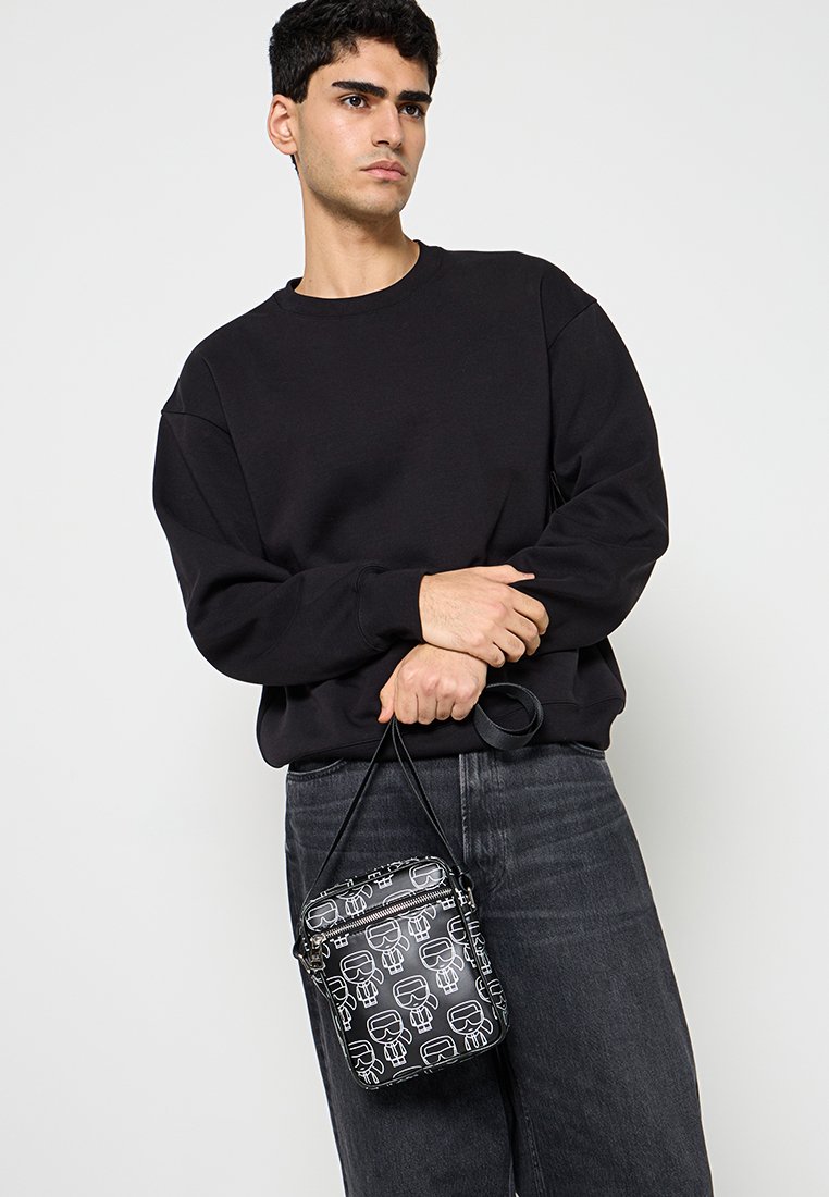 Bolso bandolera negro con patrones gráficos blancos, de forma cilíndrica, cierre con cremallera, correa adjunta, combinado con una sudadera negra y vaqueros.