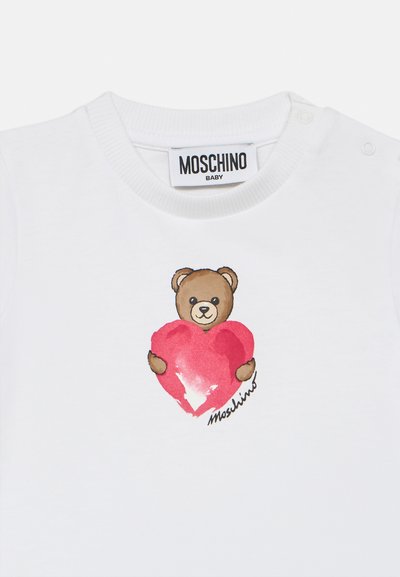 Λευκό βρεφικό μπλουζάκι με ετικέτα Moschino, που απεικονίζει μια αρκουδίτσα να κρατά μια κόκκινη καρδιά με μια μικρή υπογρα φή Moschino κάτω από αυτήν.