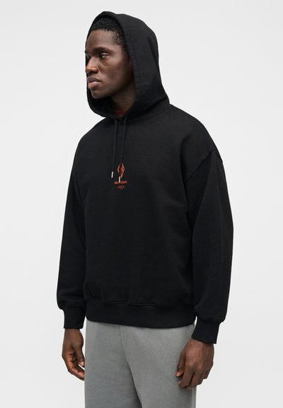 Juodas džemperis su gobtuvu, su raišteliais ir priekyje oranžinės spalvos grafika. Pagamintas iš minkštos medžiagos, turi laisvą kirpimą, ribotas rankogalius ir apačią.