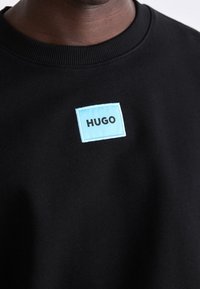 Zbliżenie na osobę w czarnej bluzie z kapturem, z małym niebieskim prostokątnym naszywką z napisem "HUGO" na klatce piersiowej.