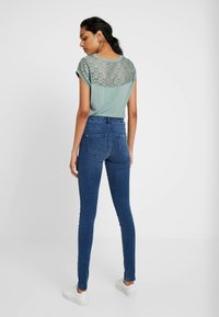Top curto azul-clarinho de mangas curtas com costas de renda estampada, combinado com jeans skinny escuros de cintura alta e sapatilhas brancas.