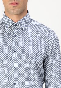 Chemise pour homme en tissu léger, à base blanche avec un motif géométrique bleu, col pointu et fermeture à boutons sur le devant.