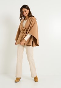 Poncho color camel con cinturón, hecho de tela suave. Presenta mangas anchas y un escote redondo, combinado con pantalones beige acampanados y botas marrones.