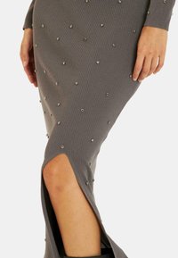 Robe grise côtelée ornée de clous métalliques, avec une fente haute sur la gauche et une silhouette ajustée à manches longues.