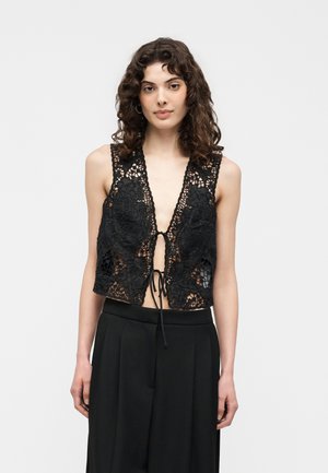 Kvinde med krøllet hår iført en ærmeløs sort blonde-top med bindebånd foran og sorte bukser med brede ben, stående mod en ensfarvet baggrund.