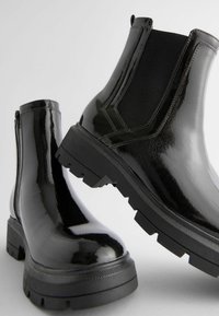Las botas Chelsea de charol negro presentan una suela gruesa, paneles laterales elásticos y un acabado liso y brillante con un ribete texturizado en contraste.