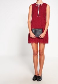 Robe sans manches en dentelle rouge avec un ruban blanc noué au cou, portant une pochette noire à la main, associée à des mocassins noirs.