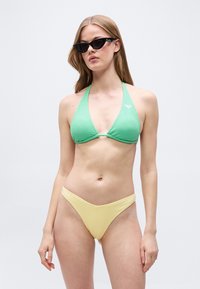 Νεαρή γυναίκα με μαύρα γυαλιά ηλίου cat-eye, πράσινο τοπ μαγιό με λαιμόκοψη halter και κίτρινο κάτω μέρος μαγιό, μπροστά σε απλό φόντο.