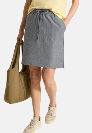 Person trägt einen grauen Nadelstreifenrock mit Kordelzug, gelbes Oberteil, beige Turnschuhe, hält eine olivgrüne gepolsterte Tragetasche.