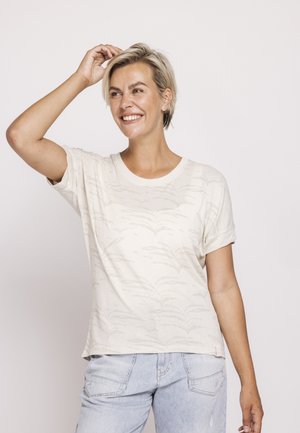 Smiling woman with short blonde hair wearing a light patterned t-shirt and light blue jeans, touching her hair with one hand.