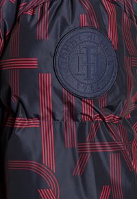 Chaqueta acolchada azul marino con patrones lineales rojos, un parche circular azul marino con el texto "Tommy Hilfiger" y el año de establecimiento 1985. Textura suave.
