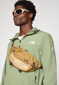 Fanny pack i tan stof med tekstureret overflade, justerbare stropper og fremtrædende branding. Båret over en grøn jakke med hætte.