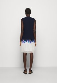 Robe sans manches avec un haut en maille bleu foncé et une jupe blanche fluide, ornée de broderies florales bleues et violettes le long de l'ourlet.