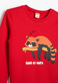 Camisa de manga larga roja con un gráfico de un mapache durmiendo, con el texto "BUENO PARA DORMIR" debajo. Tejido suave con cuello de canalé.