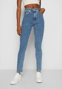 Vaqueros de mezclilla azules de cintura alta con un ajuste ajustado, que cuentan con cinco bolsillos, cremallera y detalles de costura sutiles, combinados con zapatillas blancas.