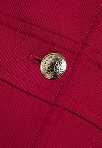 Tela roja con un tejido texturizado, que presenta un botón circular plateado embellecido con un emblema detallado y costura decorativa.