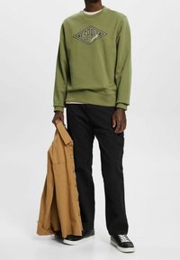 Olivgrön sweatshirt med svart logotyp, i kombination med svarta cargo-byxor. En ljusbrun skjorta med knappar hålls i vänster hand. Svarta sneakers.