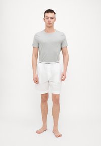 Pelēks kokvilnas T-krekls savienots ar baltām šortiem, kuriem ir melns Calvin Klein logotips jostasvietā. Modelis stāv basām kājām neitrālā vidē.