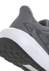 Zapatilla deportiva gris con parte superior de malla texturizada, entresuela blanca y suela de goma negra, que cuenta con un fila para el talón y una marca en la suela.