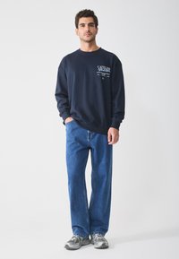 Dunkelblaues Sweatshirt mit einem Grafikdruck auf der Brust, kombiniert mit hellblauen, weit geschnittenen Jeans und grauen Sneakers. Einfaches und lässiges Outfit.