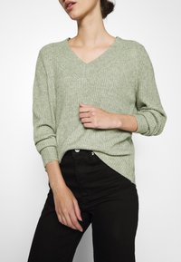 Vaaleanvihreä neulepaita, jossa on V-pääntie, ribattu tekstuuri ja kolmanneksen mittaiset hihat, yhdistettynä mustiin korkeavyötäröisiin housuihin.