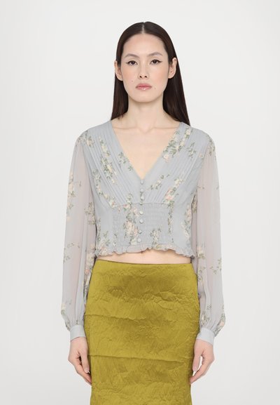 Blouse fleurie gris clair avec manches longues, décolleté en V, taille smockée et ourlet froncé. Associée à une jupe texturée vert olive.
