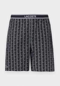 Μαύρο σορτς με γεωμετρικό μοτίβο από λευκές γραμμές, ελαστική μέση με το λογότυπο "LACOSTE" σε λευκό και μικρό έμβλημα κροκόδειλου κοντά στο τελείωμα.