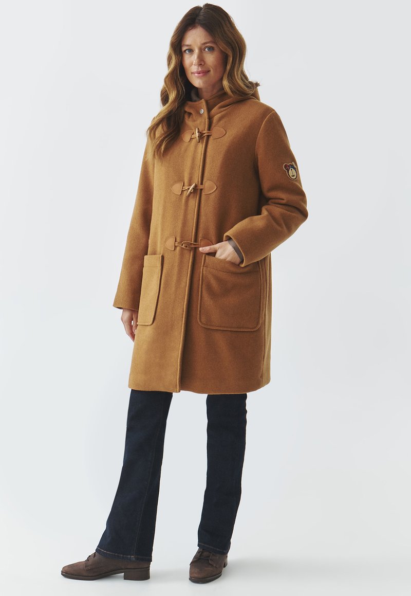 Brązowy wełniany płaszcz z zapięciem na troczki, dużymi kieszeniami i kapturem. w połączeniu z ciemnymi dżinsami i brązowymi butami. Ma naszywkę z logo na rękawie.