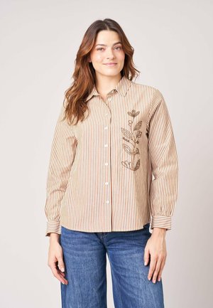 Femme portant une chemise rayée marron et blanche boutonnée avec une broderie florale sur la poitrine et un jean bleu, debout devant un fond uni.