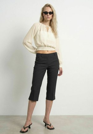 Femme portant un pantalon noir court, un chemisier crème à manches longues, des lunettes de soleil noires et des sandales à talons noires, debout devant un fond uni.