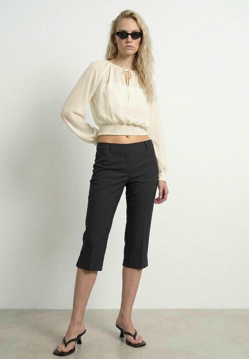 Femme portant un pantalon noir court, un chemisier crème à manches longues, des lunettes de soleil noires et des sandales à talons noires, debout devant un fond uni.
