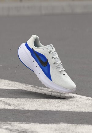 Weißer und blauer Nike-Laufschuh mit schwarzem Swoosh, schwebt über Asphalt mit weißen Fahrbahnmarkierungen und kleinen Schmutzpartikeln.