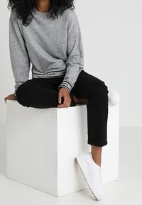 Felpa grigia a maniche lunghe con logo Levi's sull'orlo, abbinata a pantaloni neri e sneakers bianche, seduto su un cubo bianco.