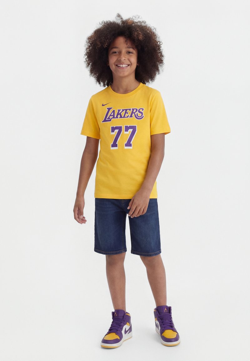 Enfant souriant aux cheveux bouclés portant un t-shirt jaune Lakers 77, un short en jean foncé et des baskets montantes violettes, jaunes et blanches, debout sur fond blanc.