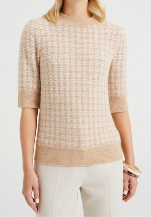 Femme portant un pull tricoté à manches courtes à carreaux beige et blanc avec des poignets et un ourlet côtelés, associé à un pantalon de couleur claire et des bracelets.