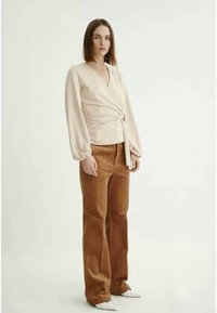 Top bege envolvente com mangas longas, tecido texturizado e nó na frente; combinado com calças largas de veludo cotelê castanhas com nervuras e sapatos brancos.