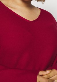 Pull à col en V, à manches longues, en tissu tricoté rouge profond. Il présente une coupe décontractée et une texture douce, avec un design d'épaule sans couture.