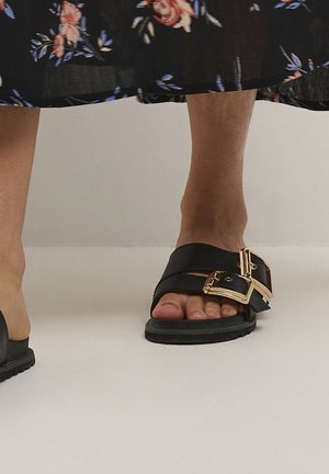 Pieds portant des sandales noires sans lacets avec de grandes boucles dorées, associées à une jupe fleurie sombre.