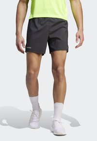 Mies, który ma na sobie czarne spodenki sportowe, białe skarpety do kostek, białe buty do biegania i neonową zieloną koszulkę sportową, stoi z lekko rozstawionymi nogami.