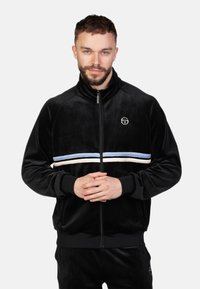 Zwarte velours zip-up jas met een hoge kraag, voorzien van witte en lichtblauwe horizontale strepen en een cirkelvormig logo op de linkerborst.
