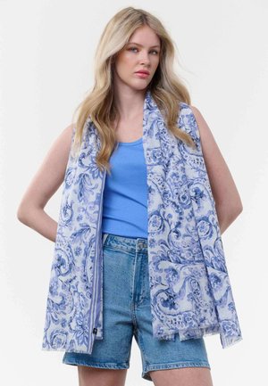 Blonde Frau trägt ein blaues Tanktop, Jeansshorts und einen hellblauen, gemusterten Schal, der über ihre Schultern gelegt ist.