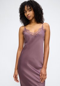 Γυναίκα με σγουρά μαλλιά που φοράει ένα μωβ σατέν φόρεμα χωρίς μανίκια, με δαντέλα στο ντεκολτέ και λεπτές τιράντες.