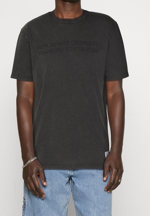 Homme portant un t-shirt noir délavé avec le texte « REPLAY NOT ORDINARY » et un jean bleu clair, debout devant un fond blanc uni.