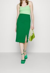 Žena kráčející v zeleném tílku, s vysokým pasem zelené sukně s rozparkem, na zelených platformách a držící světlou zelenou kabelku.